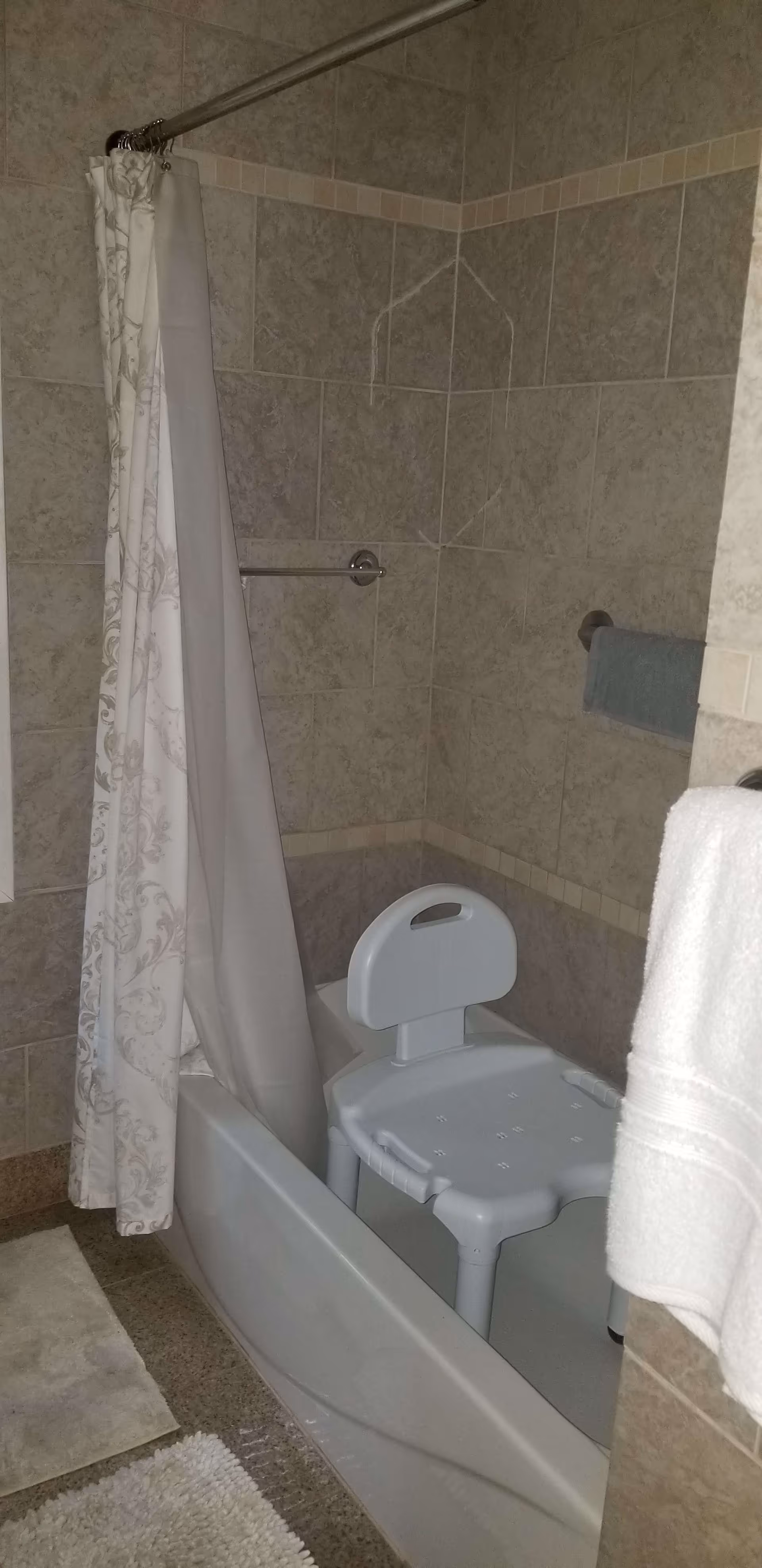 Bathroom with a bathtub that has a white shower chair inside it. The walls are tiled with beige and light brown tiles, and there is a shower curtain partially drawn to the side. A towel is hanging on a rack on the right side of the image.