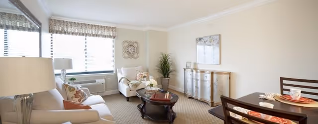 A bright and cozy living room with beige walls and carpet. The room features a beige sofa with decorative pillows, a matching armchair, a dark wooden coffee table with a book and flowers, a sideboard with a framed picture above it, and a small dining table set with plates and napkins. Large windows with blinds and valances allow natural light to fill the space.