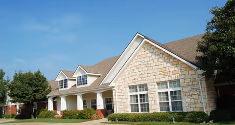 Exterior view of a single-story building with stone and siding facade, multiple windows, and a gabled roof under a clear blue sky. There are green bushes and trees around the building.