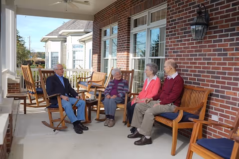 Four elderly people sitting and chatting on wooden rocking chairs and benches on a covered porch with brick walls and large windows at Maple Crest Senior Living Village.