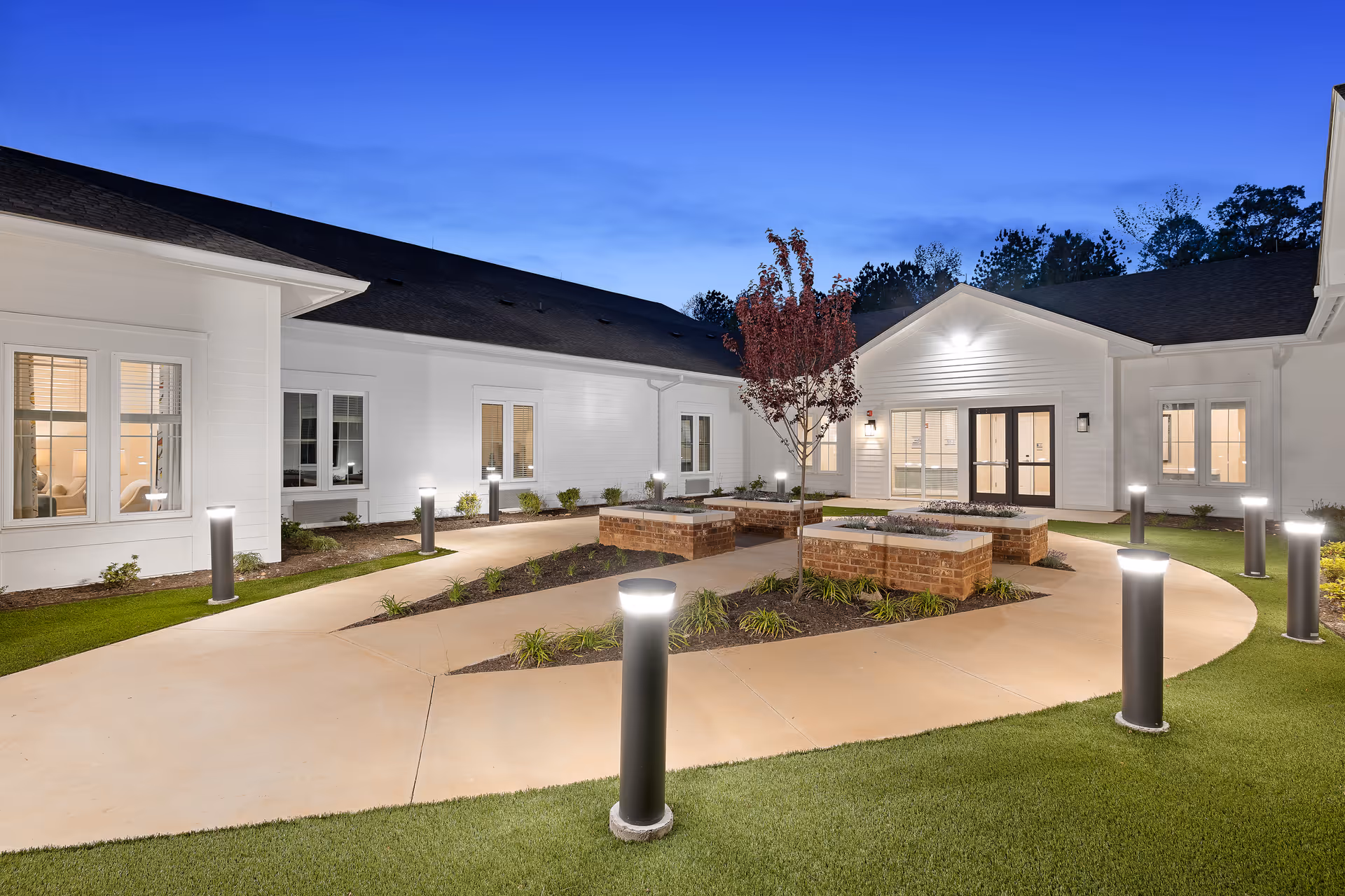 Evening view of a well-lit outdoor courtyard at The Claiborne at Brickyard Crossing, featuring a paved walkway, modern bollard lights, landscaped garden beds with a small tree, and white building walls with windows and doors.
