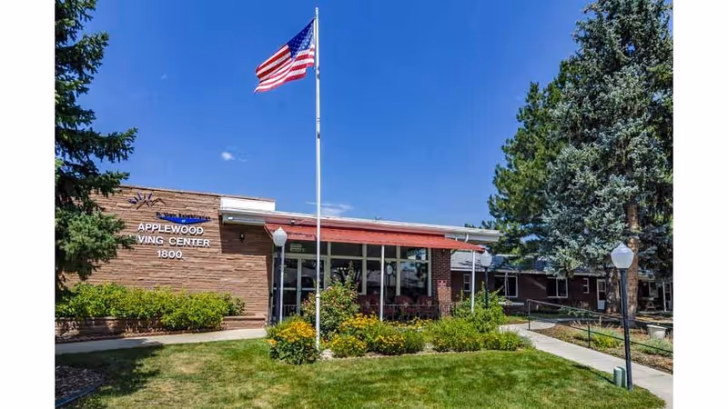 Exterior view of a single-story brick building with a sign reading 'Applewood Wing Center 1800'. There is a flagpole with an American flag in front of the building, surrounded by green grass, bushes, and trees under a clear blue sky.