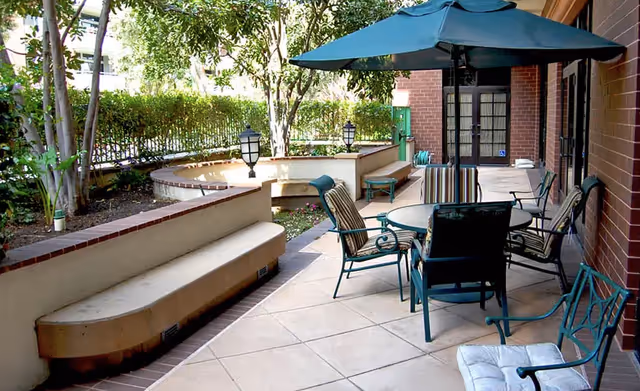 Shaded outdoor patio with a round table, umbrella, chairs, and built-in benches beside a brick building and garden.