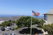 View from a higher vantage point showing a parking lot with several cars, a flagpole with an American flag, trees, and a partial view of a building on the right. In the background, there is a cityscape and a clear blue sky.