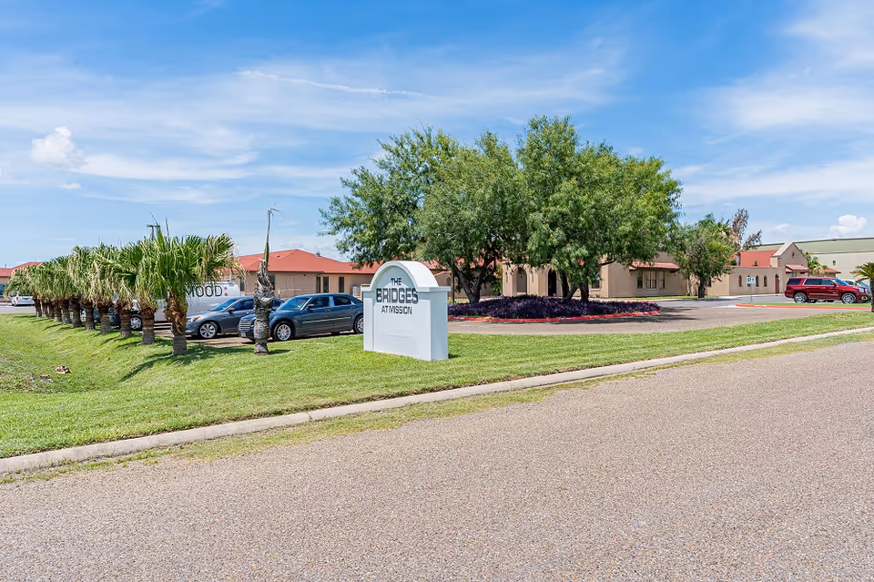 Front exterior of The Bridges at Mission Assisted Living Homes showing the entrance sign, parking lot, palm trees, and the single-story building under a blue sky.