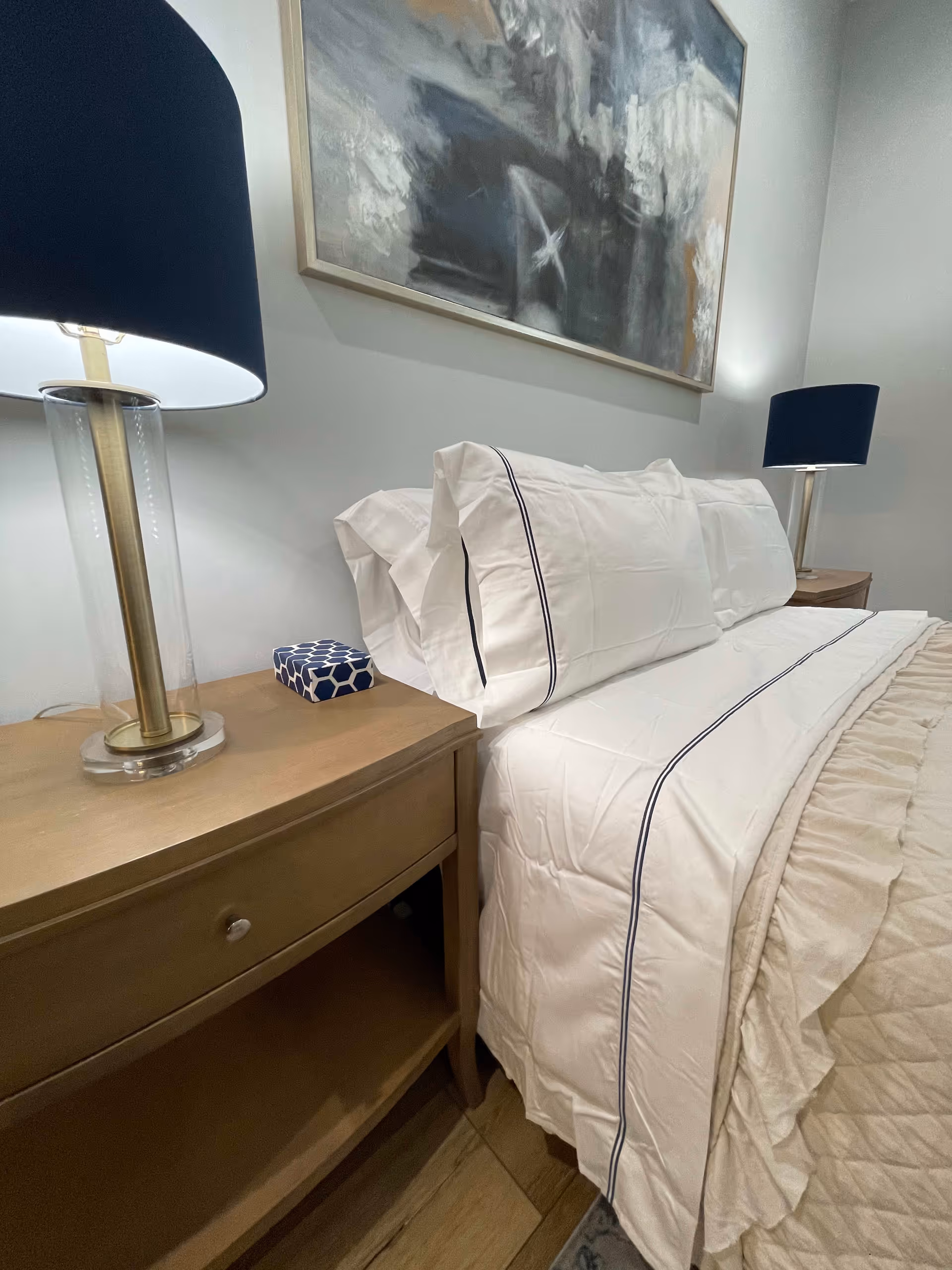 A neatly made bed with white sheets and pillowcases featuring dark piping, flanked by two wooden nightstands each with a lamp that has a clear base and a dark blue lampshade. Above the bed hangs an abstract painting in muted tones.