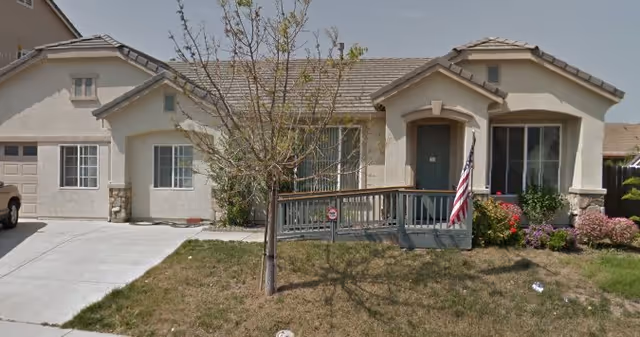 Single-story residential building with a beige exterior, a tiled roof, and a front porch with a ramp. There is an American flag displayed near the entrance, a small tree in the front yard, and some bushes and flowers near the porch. A driveway is visible on the left side of the house.