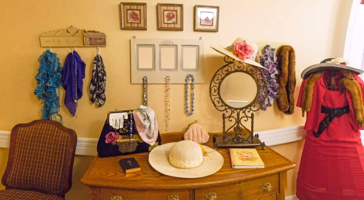 A vintage-style vanity area with a wooden dresser holding a large white hat, a small book, a purse with gloves and a scarf, and a decorative round mirror with an ornate metal frame. On the wall above the dresser are three framed floral pictures, a rack with hanging scarves and necklaces, and a mannequin dressed in a red dress with fur stole and a hat.