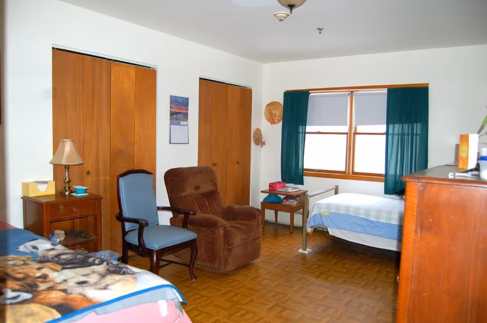 A simple bedroom with two beds, one with a dog-themed blanket and the other with a blue and white checkered blanket. The room has wooden parquet flooring, two wooden closets with sliding doors, a window with green curtains, a brown recliner chair, a blue cushioned chair, a wooden nightstand with a lamp and tissue box, and a wooden dresser.