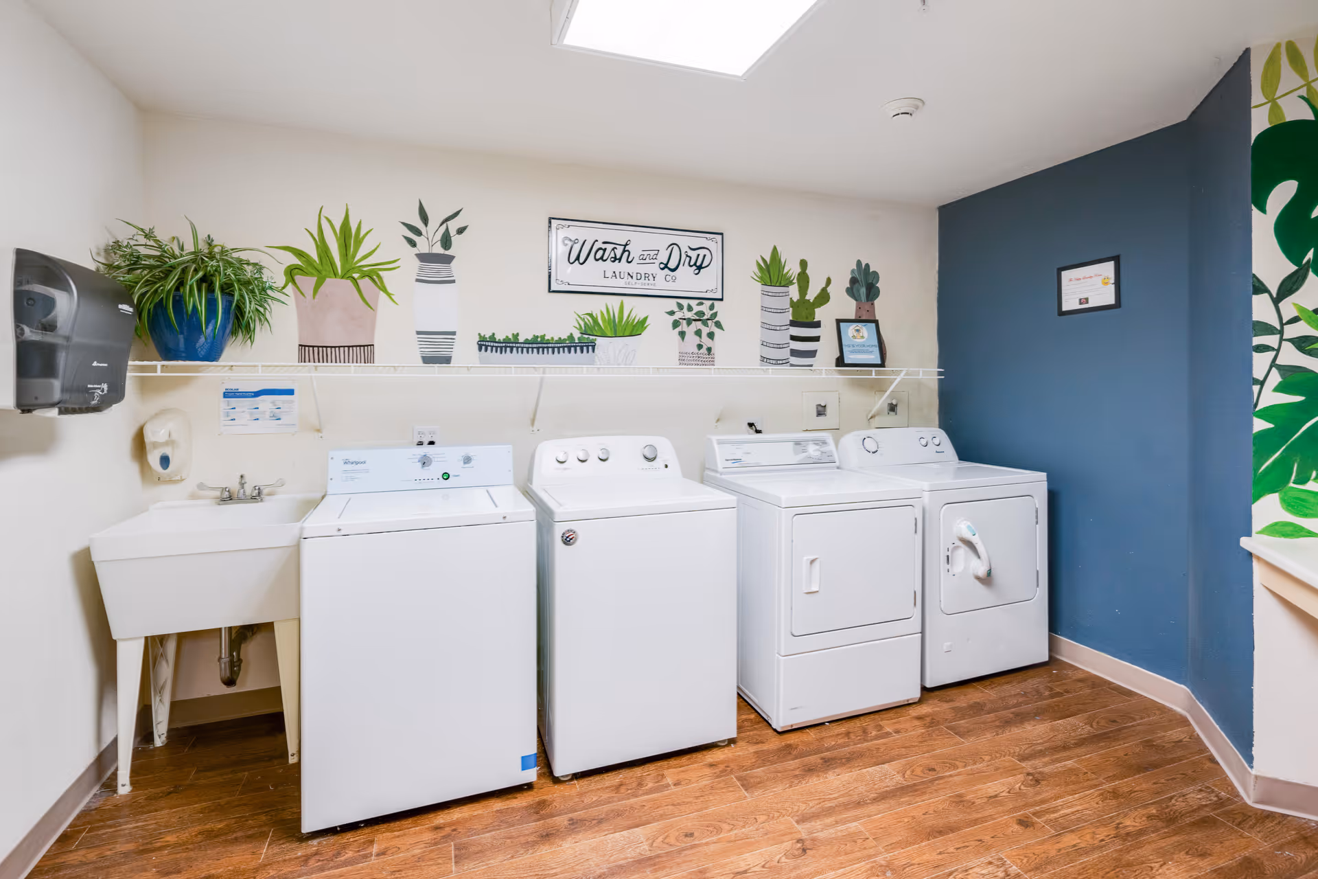 Laundry room with four white laundry machines including washers and dryers, a utility sink on the left, a paper towel dispenser and soap dispenser mounted on the wall, and decorative plant decals on the wall above the machines. The floor is wood, and one wall is painted dark blue.