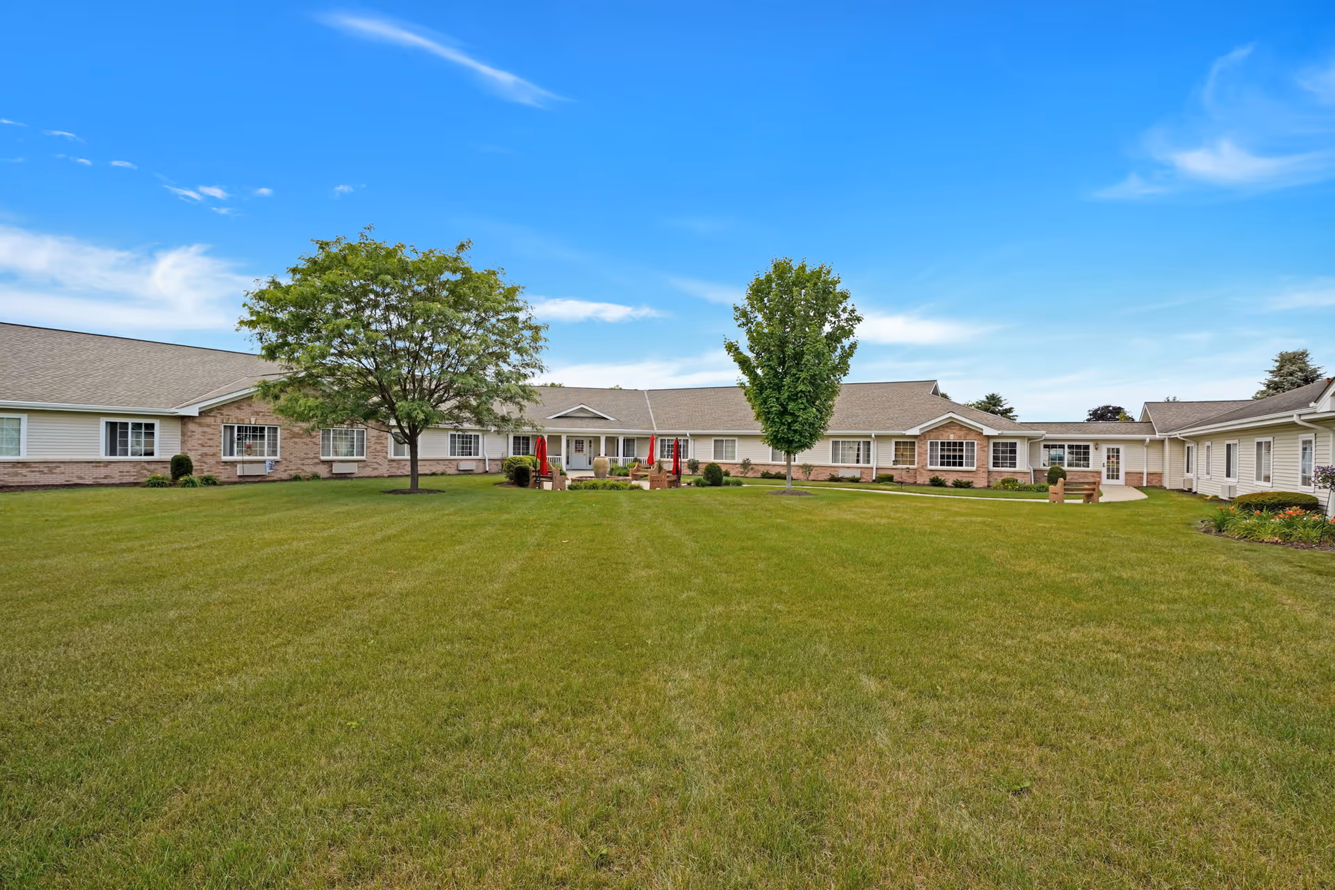 Single-story senior living building surrounding a large grassy courtyard with trees and outdoor seating under a blue sky.