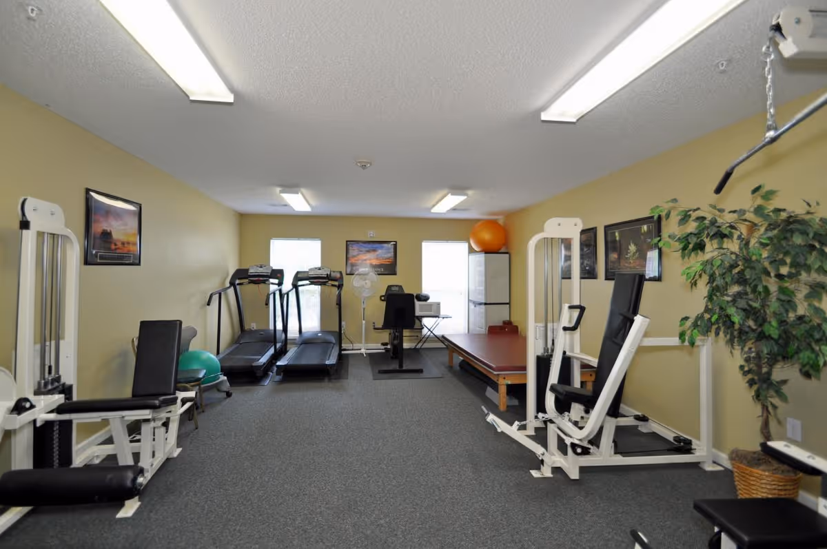 A small fitness room with exercise equipment including two treadmills, weight machines, a stationary bike, an exercise ball, and a padded exercise table. The room has beige walls, gray carpet, fluorescent ceiling lights, framed pictures on the walls, and a potted plant in the corner.