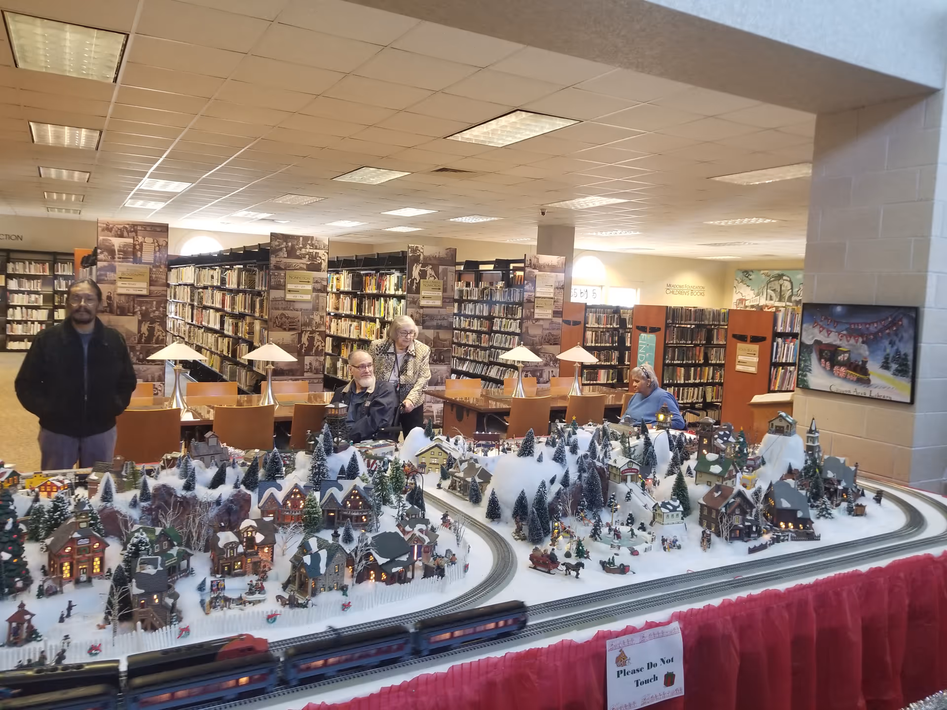 A library interior with bookshelves and tables with lamps. In the foreground, there is a detailed miniature winter village display with small houses, trees, and a train set on snowy terrain. Three elderly people are visible near the display, one standing and two seated. The library has a warm, well-lit atmosphere.