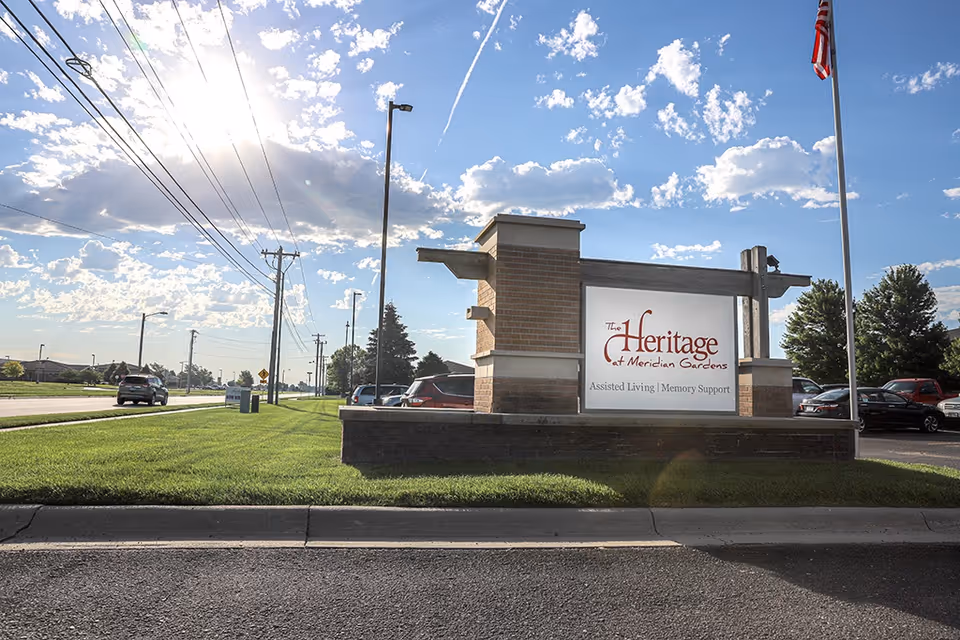Outdoor view of a street with a large sign for The Heritage at Meridian Gardens, an assisted living and memory support facility. The sign is made of brick and wood, with a flagpole and American flag to the right. Several cars are parked behind the sign, and the sky is partly cloudy with the sun shining brightly.