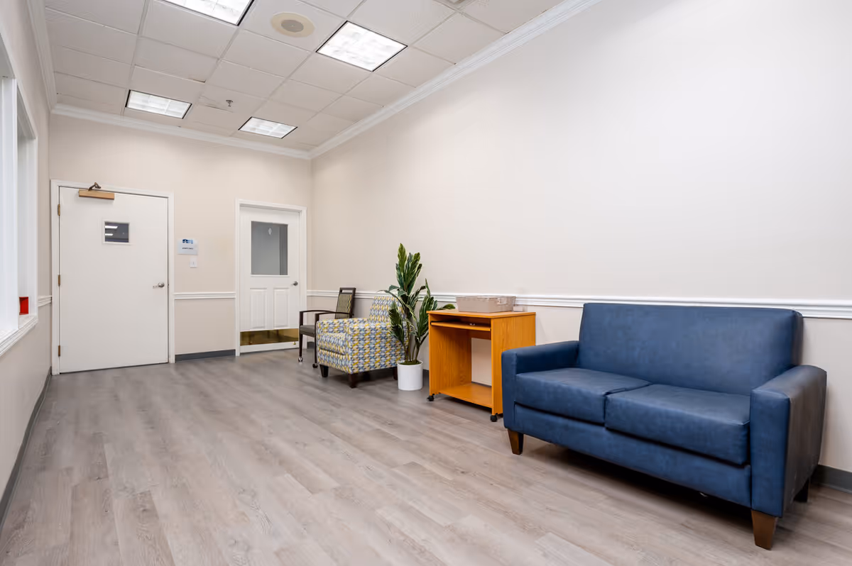 A waiting area with a blue two-seater sofa, a patterned armchair, a wooden side table with a basket on top, and a potted plant. The room has light-colored walls, a gray wood floor, and two white doors. The ceiling has recessed lighting panels.