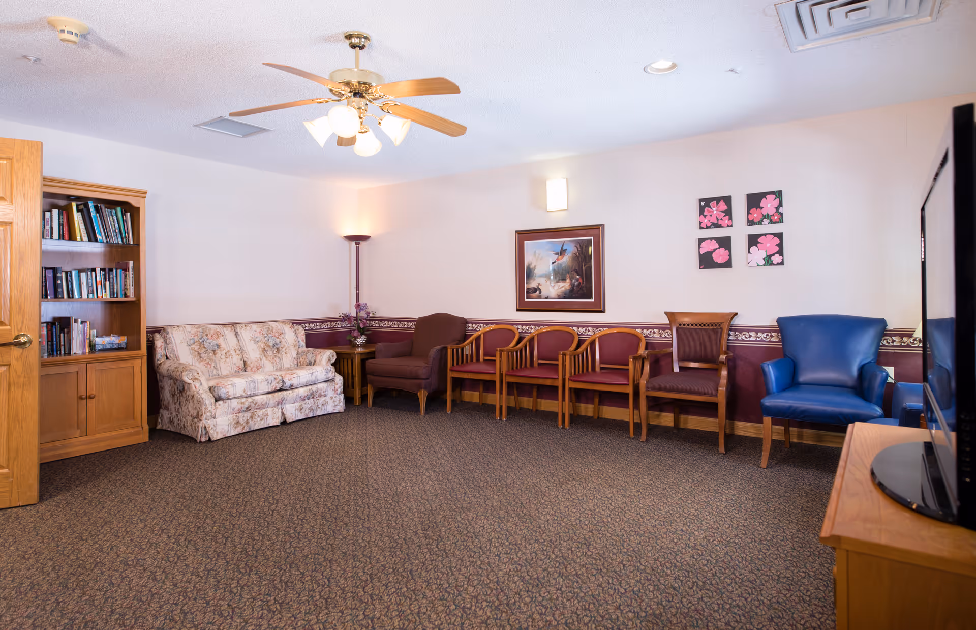 A communal sitting room with a floral sofa, a row of wooden chairs, bookcase, blue armchair, TV and a ceiling fan.