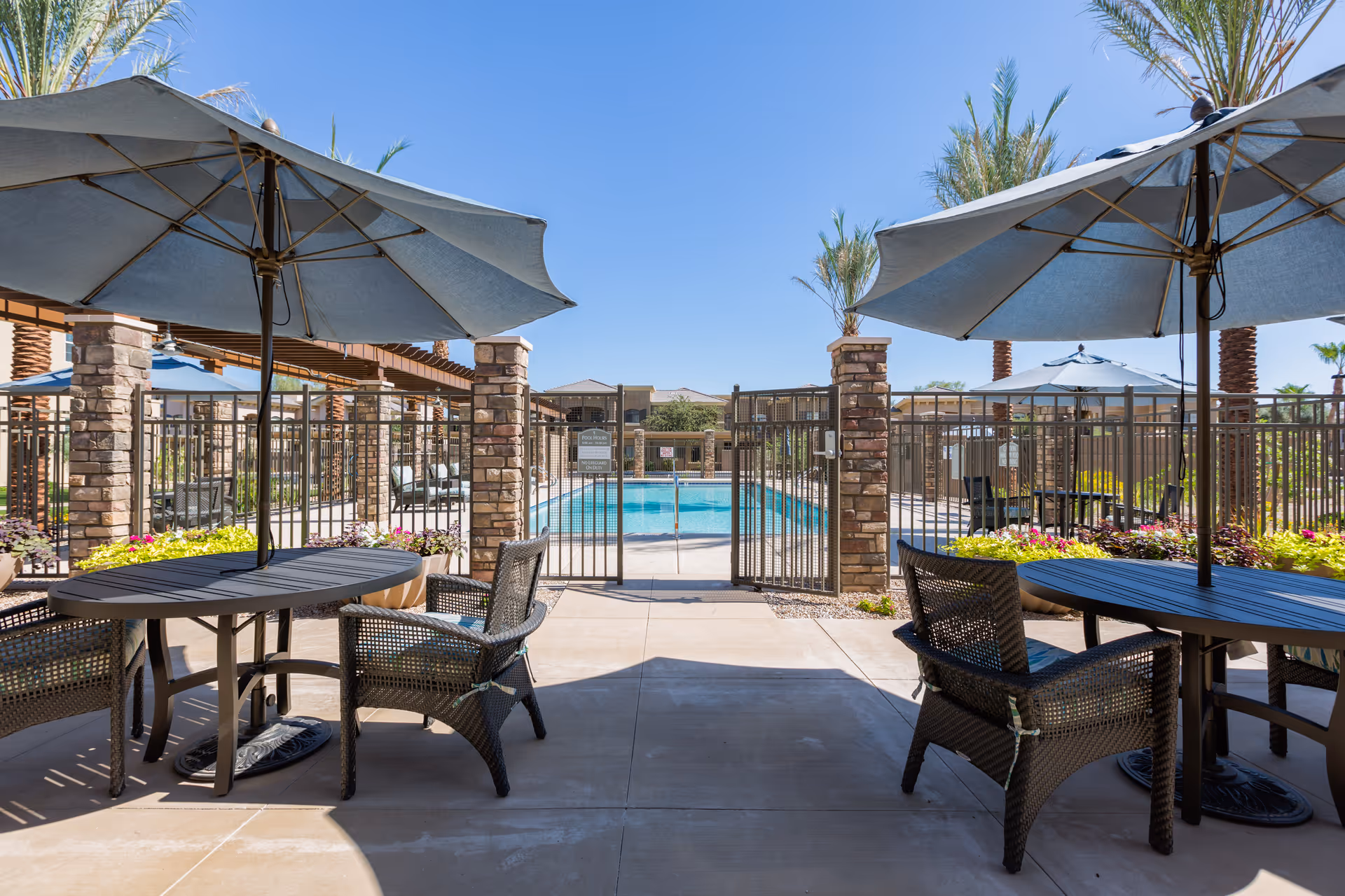 Outdoor seating area with round tables and umbrellas next to a fenced swimming pool, surrounded by palm trees and landscaping under a clear blue sky.