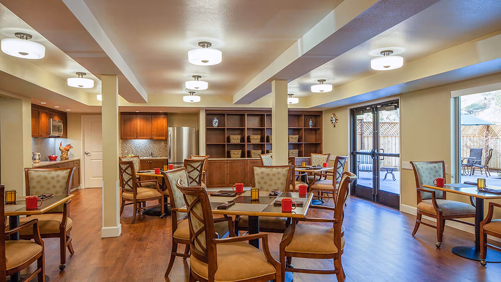 A well-lit dining area with multiple wooden tables and cushioned chairs arranged neatly. Each table has placemats, red mugs, and small decorative candle holders. The room features wooden flooring, a kitchenette with cabinets, a refrigerator, and a microwave on the left side. Large windows and glass doors on the right side provide a view of an outdoor patio with seating. The ceiling has multiple modern light fixtures.