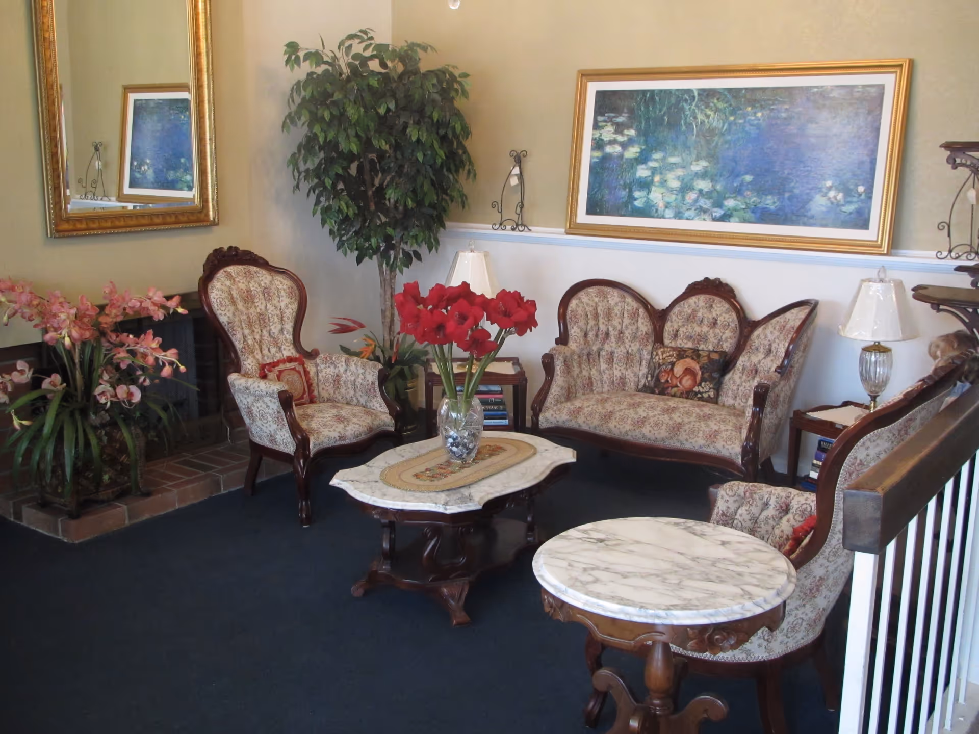 A cozy living room with vintage floral upholstered chairs and a loveseat arranged around two wooden tables with marble tops. A vase with red flowers is on one table. The room features a large framed painting of water lilies on the wall, a tall green plant in the corner, a mirror, and a fireplace with pink flowers on the hearth.