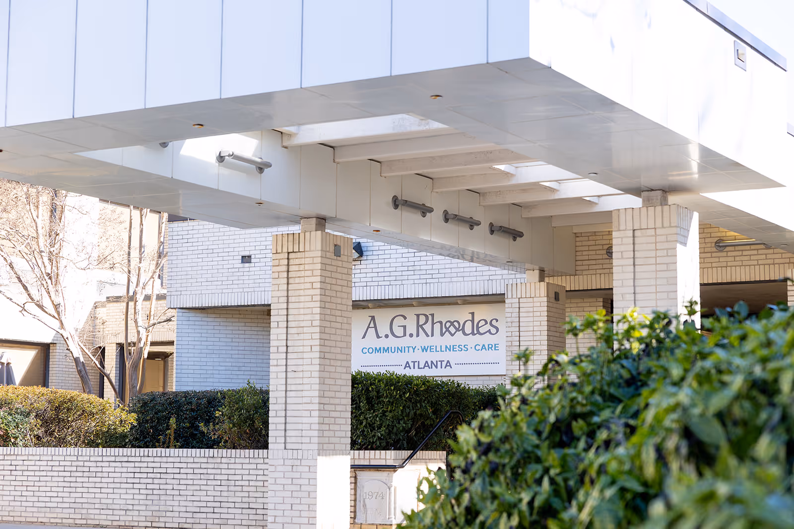 Exterior view of the A.G. Rhodes Atlanta facility showing a white brick building with a covered entrance supported by columns and greenery in the foreground.