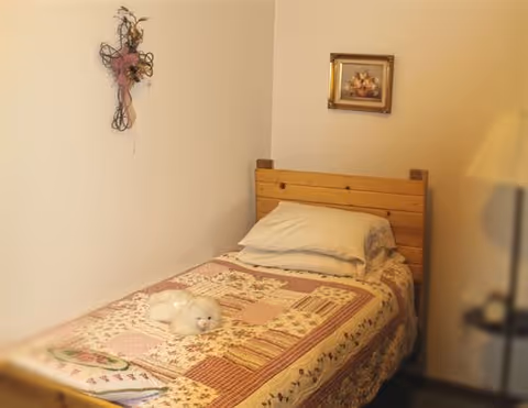 A small bedroom with a single wooden bed covered with a patchwork quilt. A white stuffed animal is placed on the bed. On the wall above the bed, there is a decorative cross with flowers and a small framed picture. A lamp is partially visible on the right side.