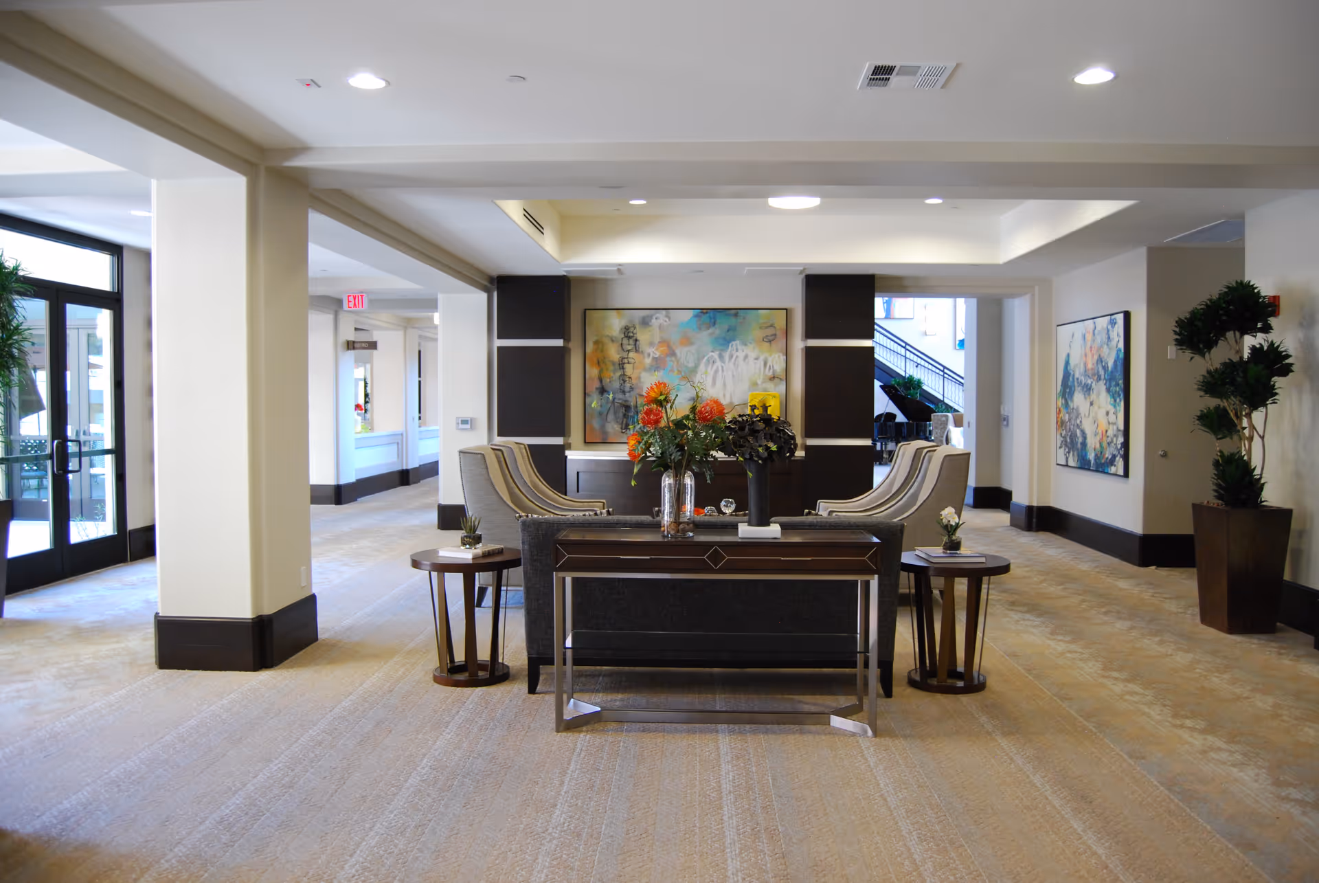 A spacious and well-lit interior common area with beige walls and carpeted floors. The room features a seating arrangement with a dark sofa and four beige armchairs around a central table adorned with flower vases. There are side tables with small plants on either side of the sofa. The walls are decorated with colorful abstract paintings, and large windows and glass doors allow natural light to enter. Potted plants are placed in the corners, and a staircase is visible in the background.