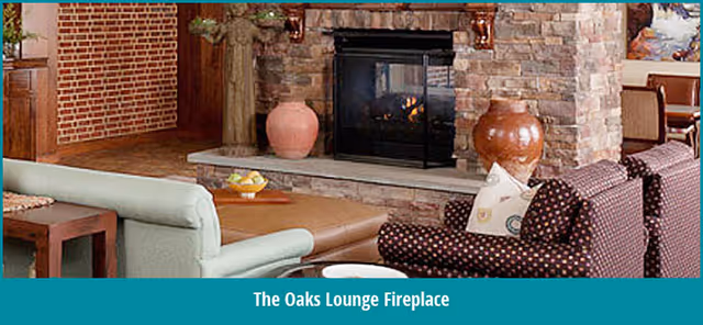 Cozy lounge area with a stone fireplace, two patterned armchairs, a light green sofa, a wooden side table, and decorative vases on the fireplace hearth.