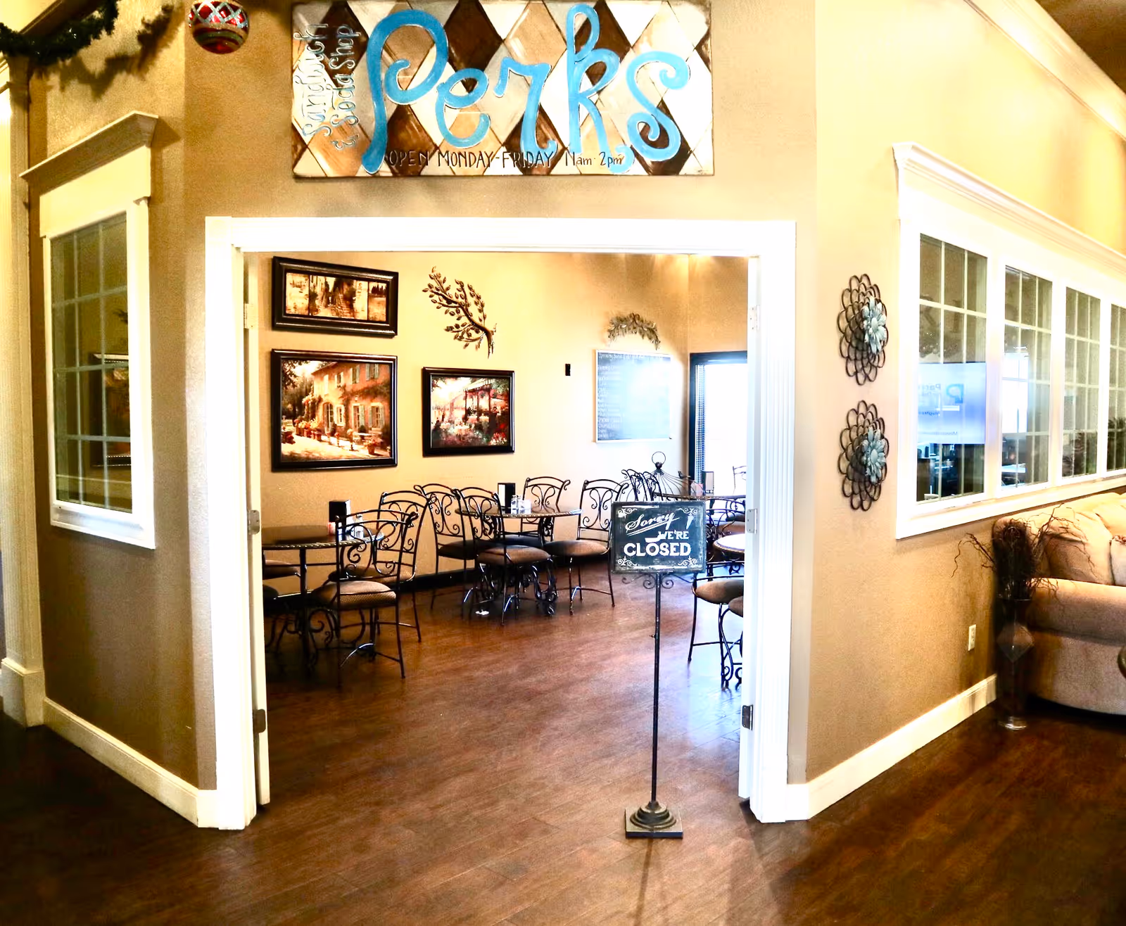Interior view of a dining area with several metal chairs and tables. The room has framed artwork on the walls and decorative wall hangings. A sign in the foreground reads 'Sorry We're Closed.' Above the doorway is a colorful sign that says 'Perks' with hours listed as Monday to Friday 11am to 2pm. The area outside the dining room has a beige wall, windows, and a couch with a plant beside it.