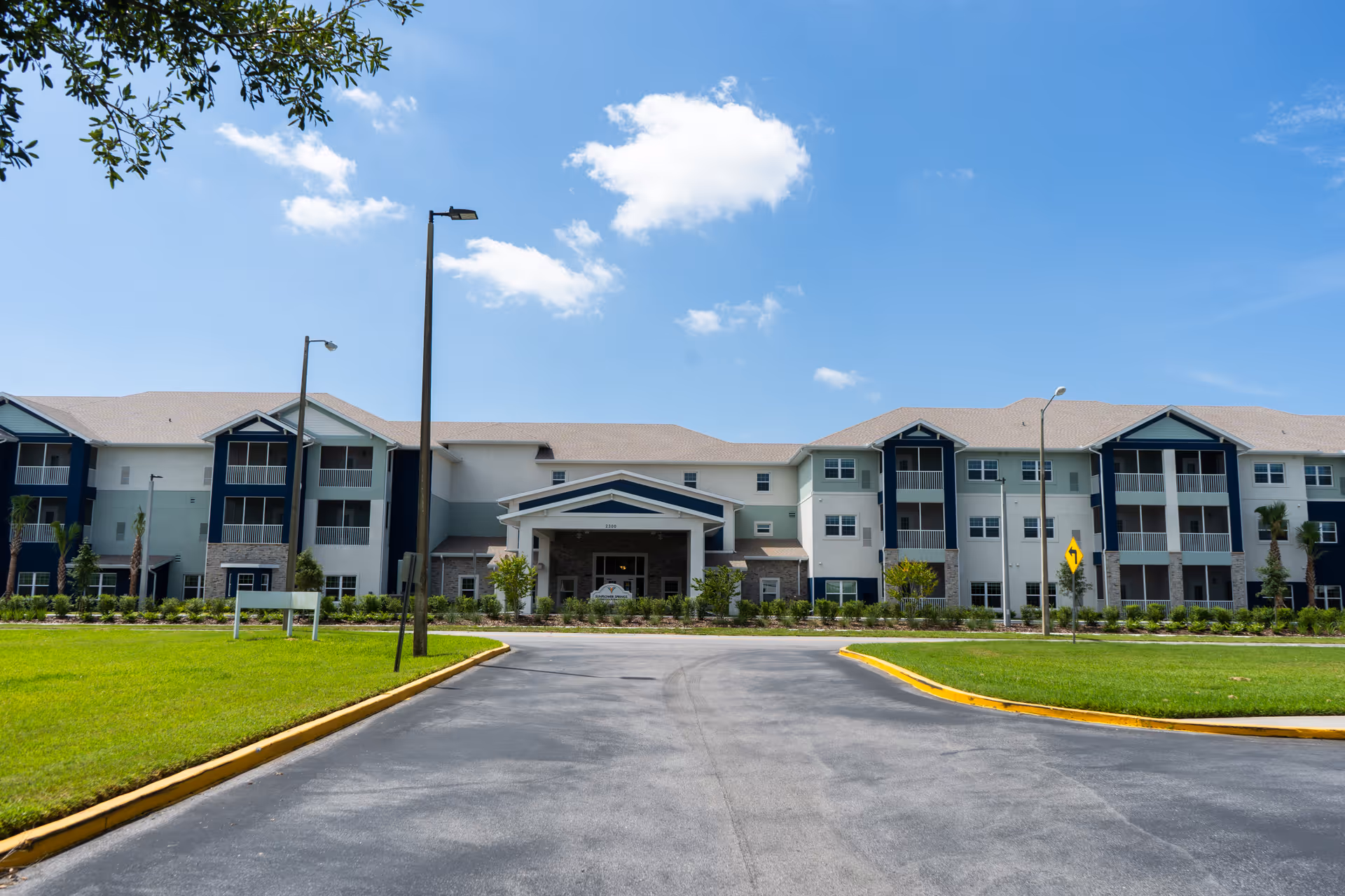 Front exterior of a three-story senior living building with a covered entrance, balconies, and a driveway under a blue sky.