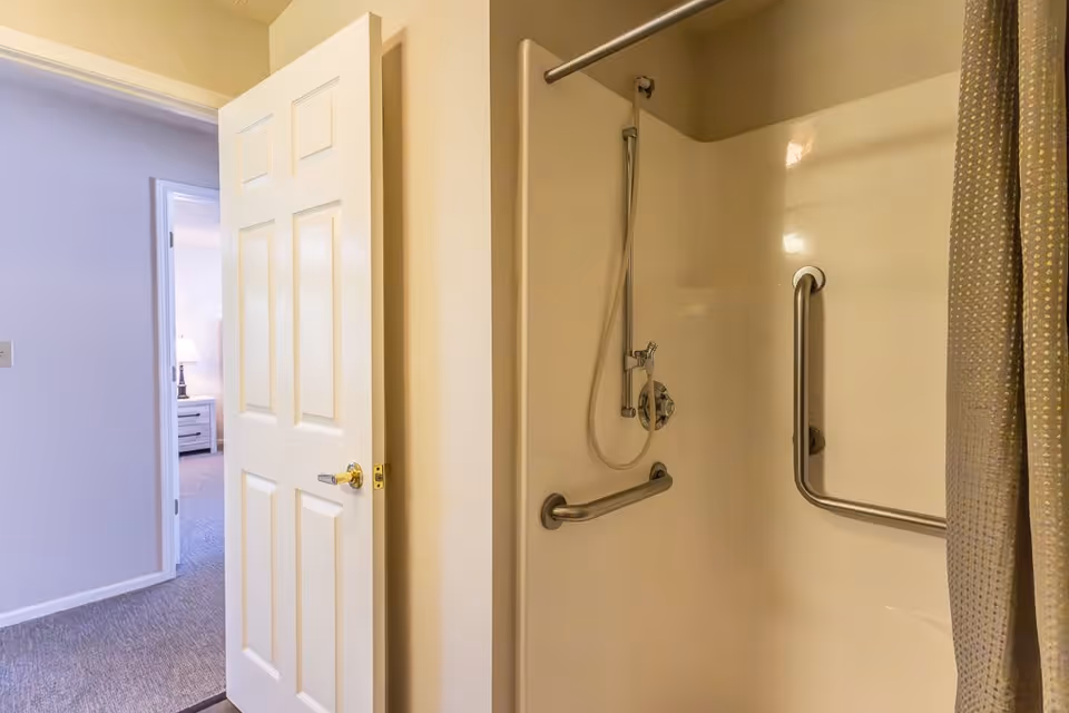View of a bathroom shower area with grab bars and a handheld showerhead, partially covered by a shower curtain. The bathroom door is open, revealing a glimpse of a bedroom with a nightstand and lamp.