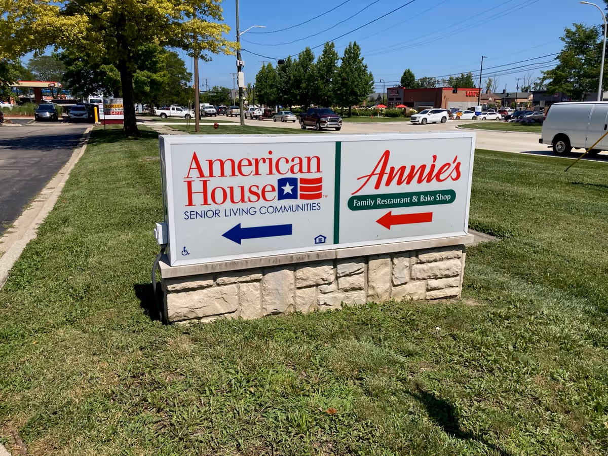 Outdoor image of a sign on a grassy area showing directions to American House Senior Living Communities with a blue arrow pointing left, and Annie's Family Restaurant & Bake Shop with a red arrow pointing right. The background includes a street, parked cars, trees, and buildings under a clear blue sky.