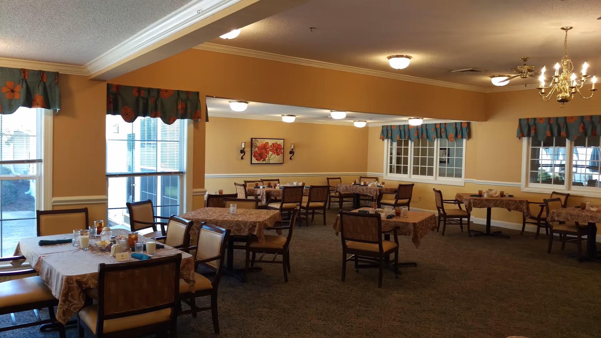 A dining room with multiple tables covered with patterned tablecloths and set with glasses, cups, and napkins. The room has beige walls, large windows with blue and orange floral valances, and a chandelier hanging from the ceiling. Chairs with cushioned seats surround the tables.