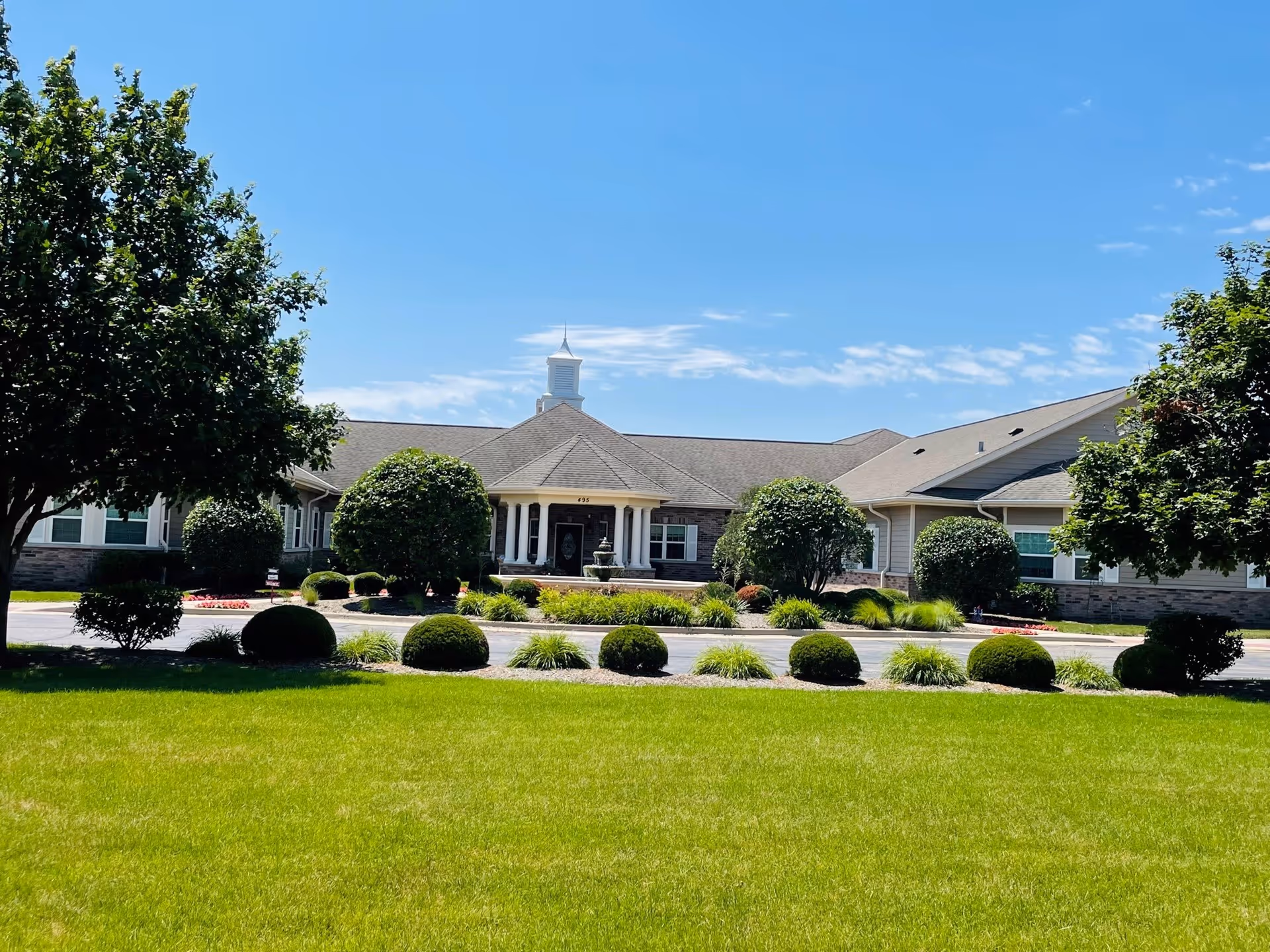 Front exterior view of Encore Memory Care at Crystal Lake building with a well-maintained lawn, neatly trimmed bushes, and trees under a clear blue sky.