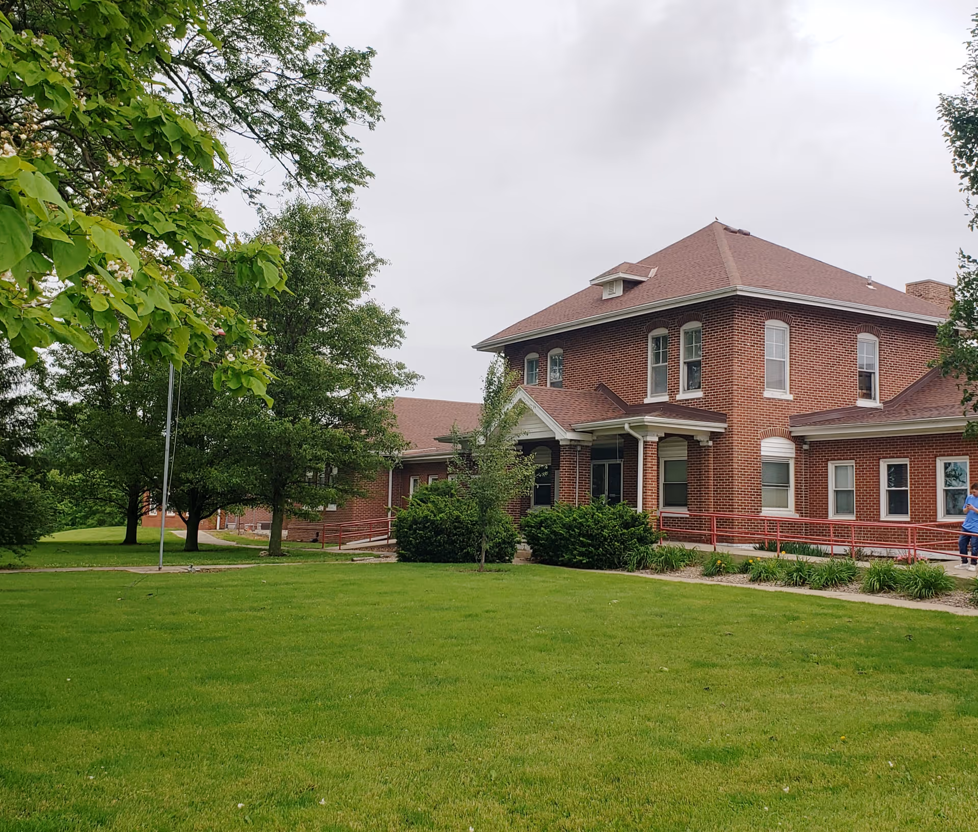 Two-story red-brick care facility with a front porch, red accessibility ramp, green lawn and trees under an overcast sky.