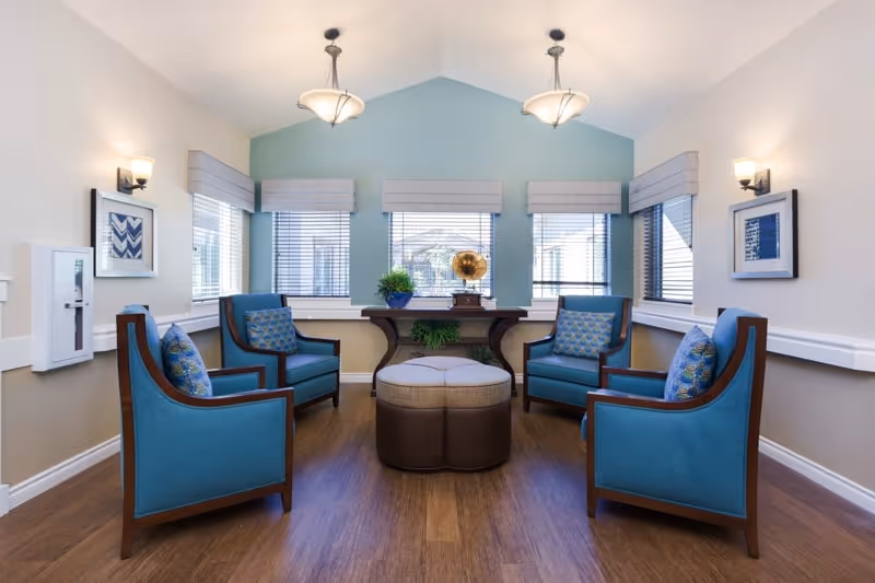 A cozy sitting area with four blue upholstered armchairs arranged around a round ottoman in the center. The room has light-colored walls with a blue accent wall featuring three windows with blinds. A wooden console table beneath the windows holds a vintage gramophone and a potted plant. The floor is wooden, and the ceiling has two hanging light fixtures. Framed artwork is displayed on the walls.
