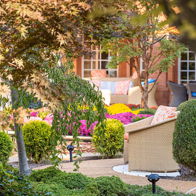 A vibrant outdoor garden area with colorful flowers, green shrubs, and trees. There are comfortable wicker chairs with patterned cushions arranged on a paved patio in front of a brick building with large windows.