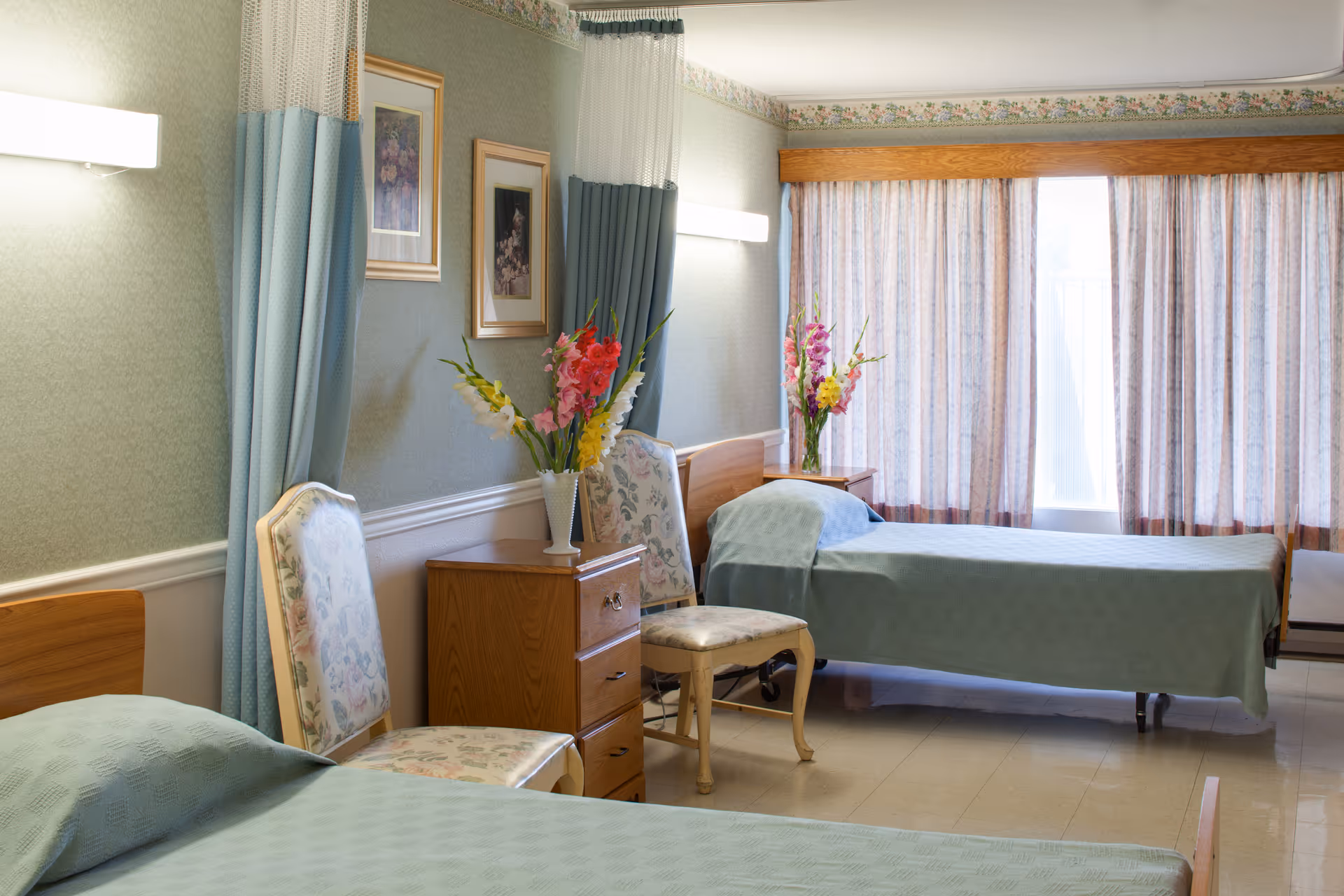 A shared bedroom in a senior living facility with two single beds covered in light blue bedspreads. Between the beds are two wooden nightstands, each with a vase of colorful flowers. Two upholstered chairs with floral patterns are placed near the beds. The room has green wallpaper, framed pictures on the wall, and large windows with sheer curtains allowing natural light to enter.
