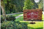 A brick sign for Courtyard Assisted Living Community situated on a grassy area with trees and shrubs surrounding it, and a paved pathway visible in the background.