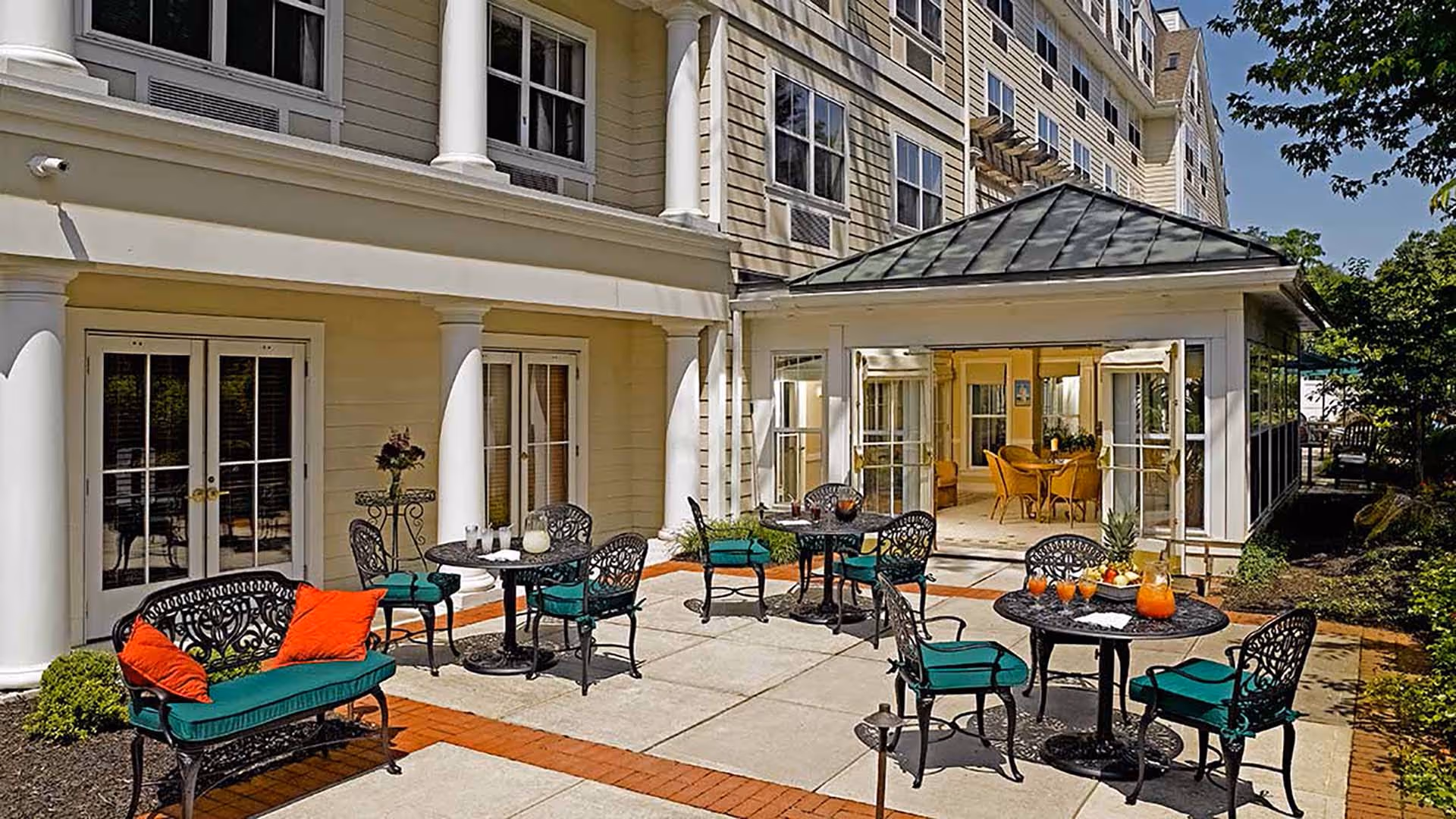 Outdoor patio area at a senior living facility with black wrought iron tables and chairs featuring green cushions and orange pillows. The patio is adjacent to a beige building with white columns and large windows. One table has a pitcher of orange juice and glasses, and there are plants and trees around the patio.