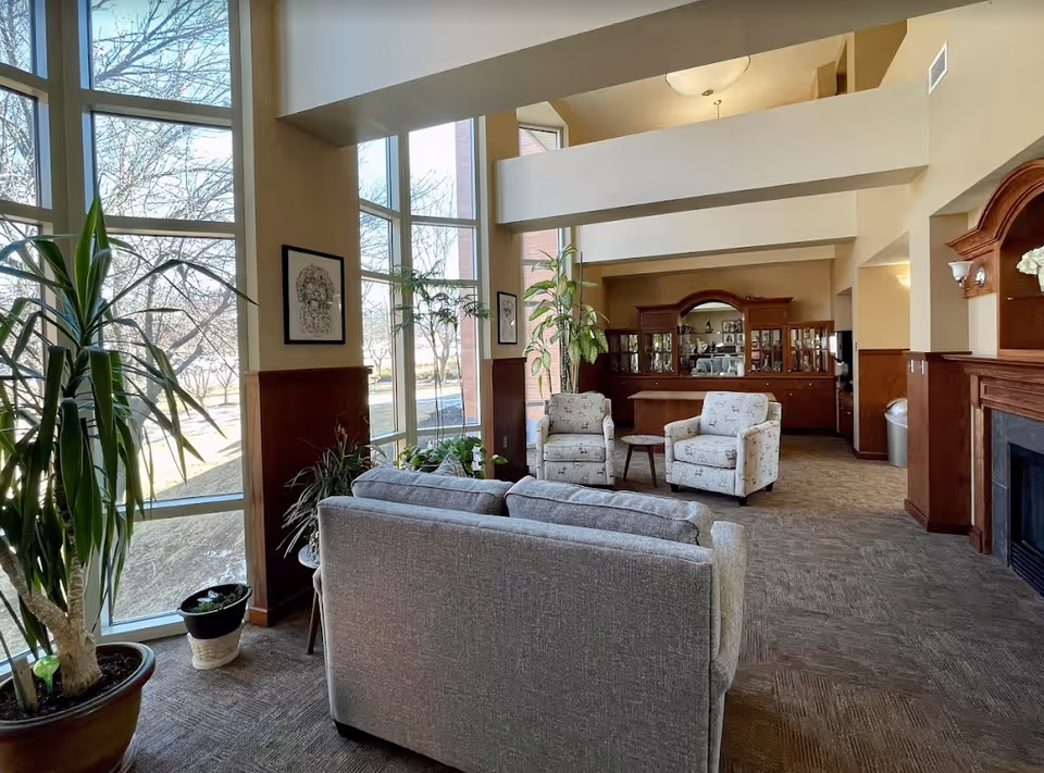 A bright and spacious living room area with large floor-to-ceiling windows letting in natural light. The room features a gray sofa, two patterned armchairs, a small round wooden table, several potted plants, and a wooden cabinet with glass doors in the background. The walls are painted beige with wood paneling halfway up, and there is a fireplace on the right side.