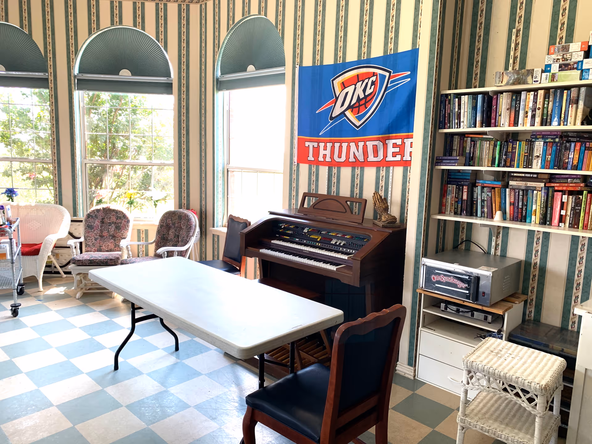 A bright room with three large windows covered by green half-circle blinds. The room features a white folding table surrounded by chairs, a wooden organ against the wall with an Oklahoma City Thunder flag hanging above it, a bookshelf filled with books and board games, and a small white wicker table. The floor has a checkered pattern with light blue and white tiles, and the walls are decorated with striped wallpaper.