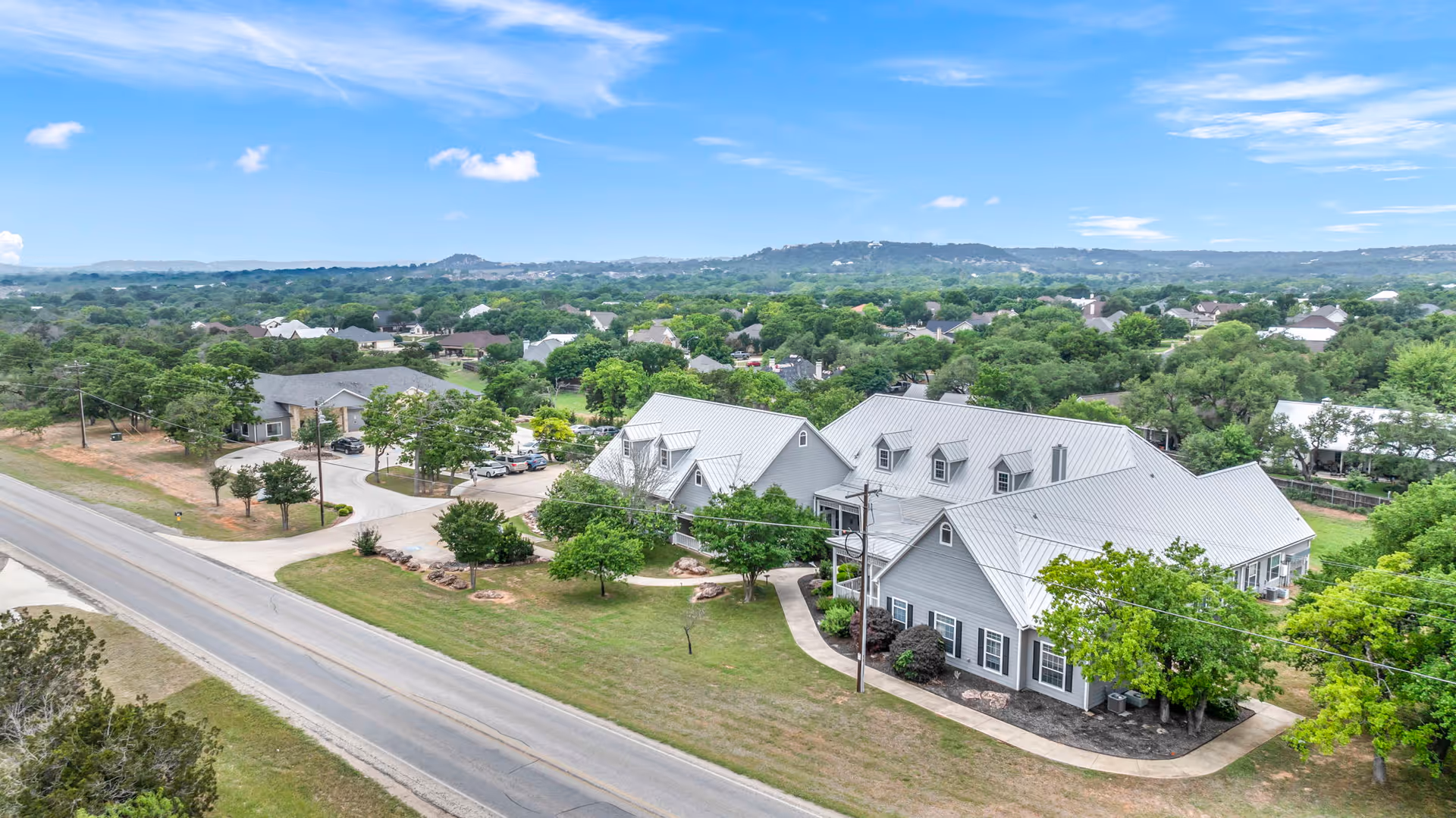 Aerial view of gray-roofed senior living buildings with lawns, trees and a road in front.