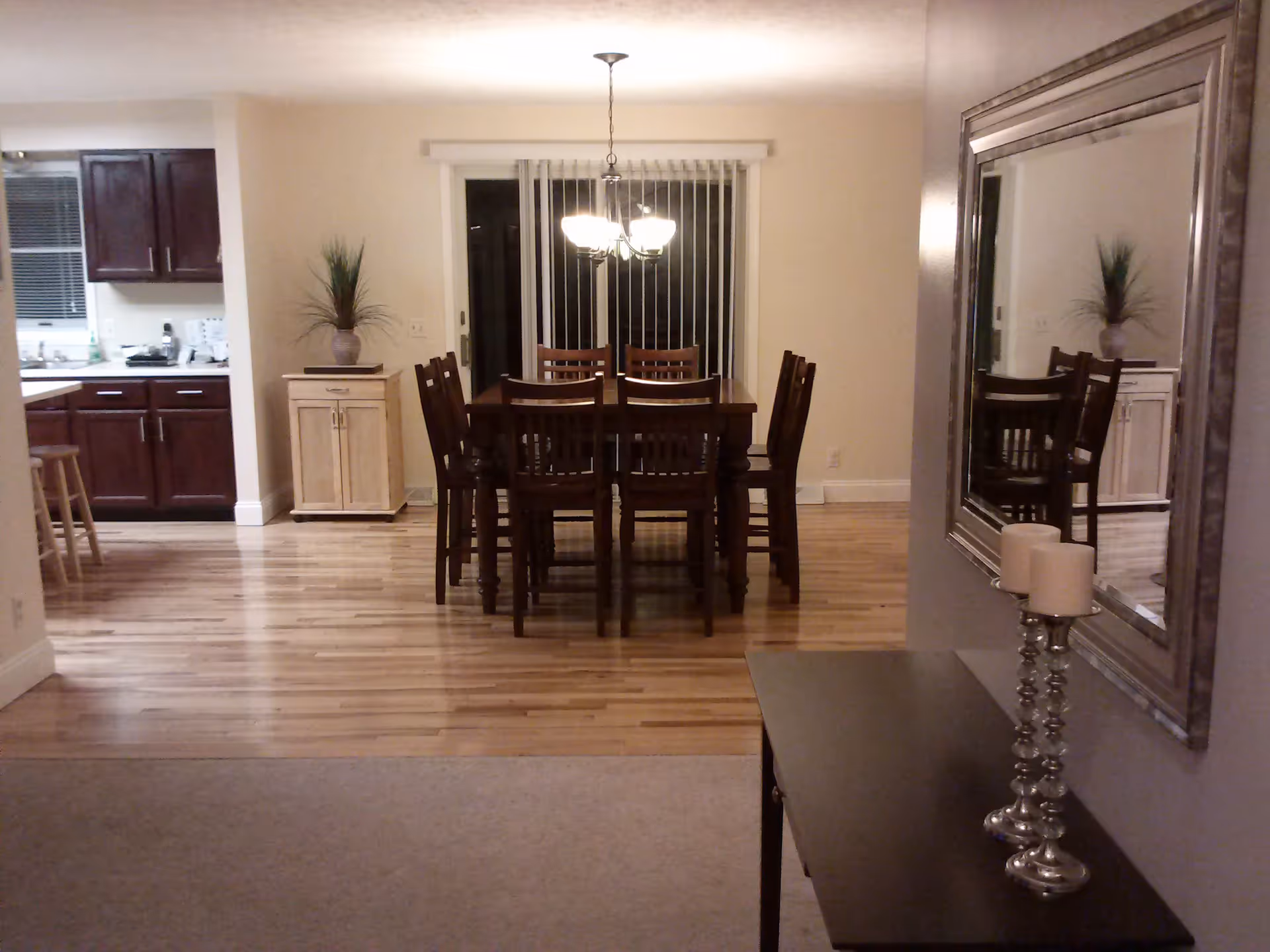 Open-plan dining area with a wooden table and chairs under a chandelier, adjacent to a kitchen and reflected in a large wall mirror.