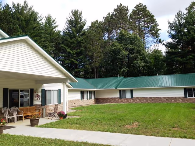 Exterior view of a single-story building with a green metal roof and white siding with brick accents. There is a covered porch area with chairs, a bench, and potted plants. The building is surrounded by a grassy lawn and tall trees in the background under a partly cloudy sky.