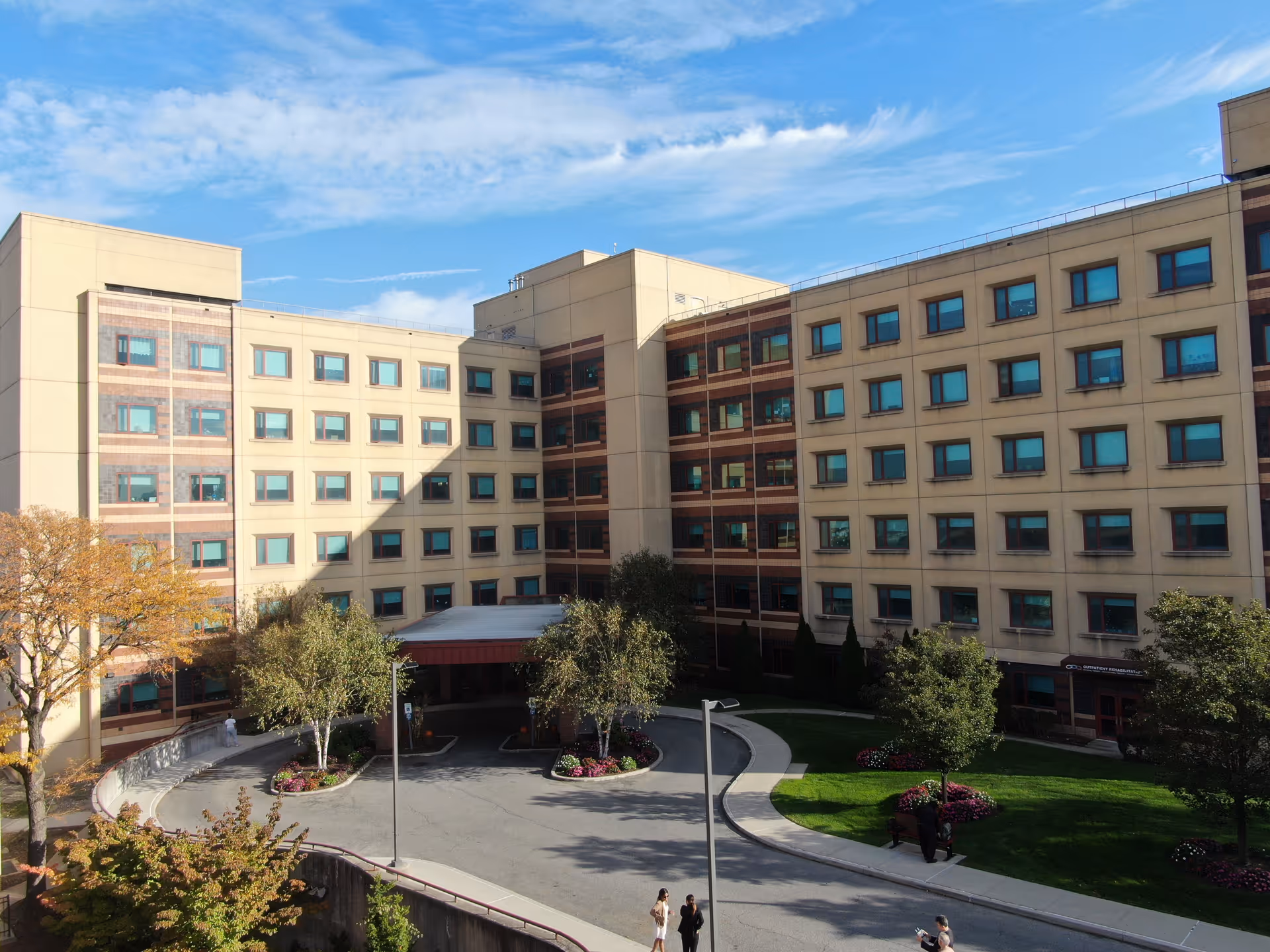 Exterior view of a multi-story building with numerous windows, surrounded by trees and landscaped greenery under a partly cloudy sky. A driveway curves in front of the building entrance where a few people are walking.