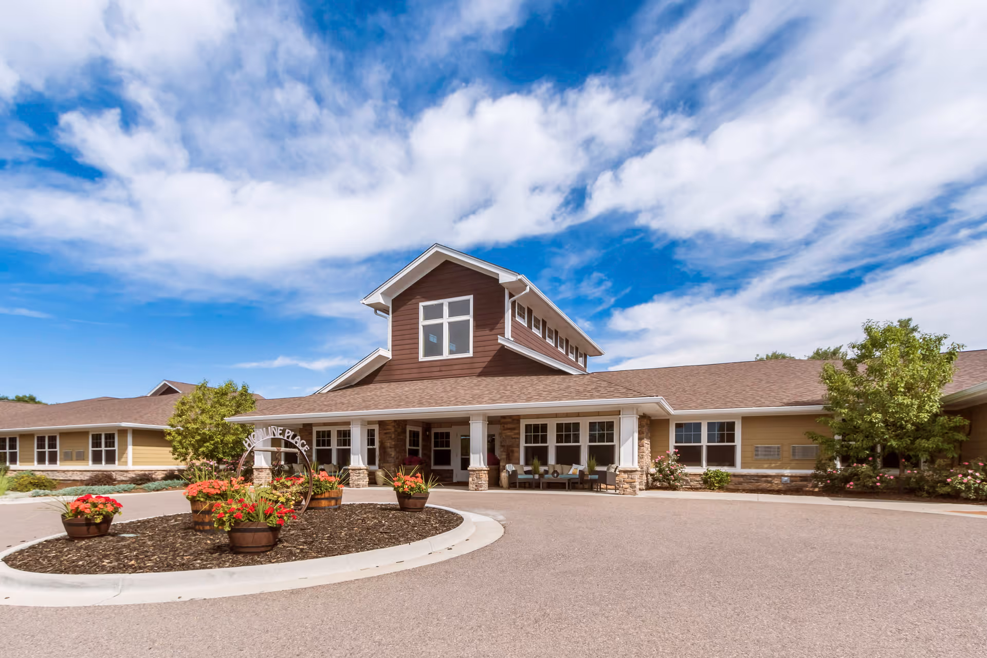 Front exterior view of Highline Place Memory Care facility with a circular driveway, flower pots with red flowers, and a clear blue sky with some clouds.