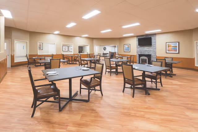 A spacious dining room with multiple tables and chairs arranged neatly. Each table has place settings with plates and napkins. The room features wood flooring, beige walls with wainscoting, framed artwork, a wall-mounted TV above a stone fireplace, and bright ceiling lights.