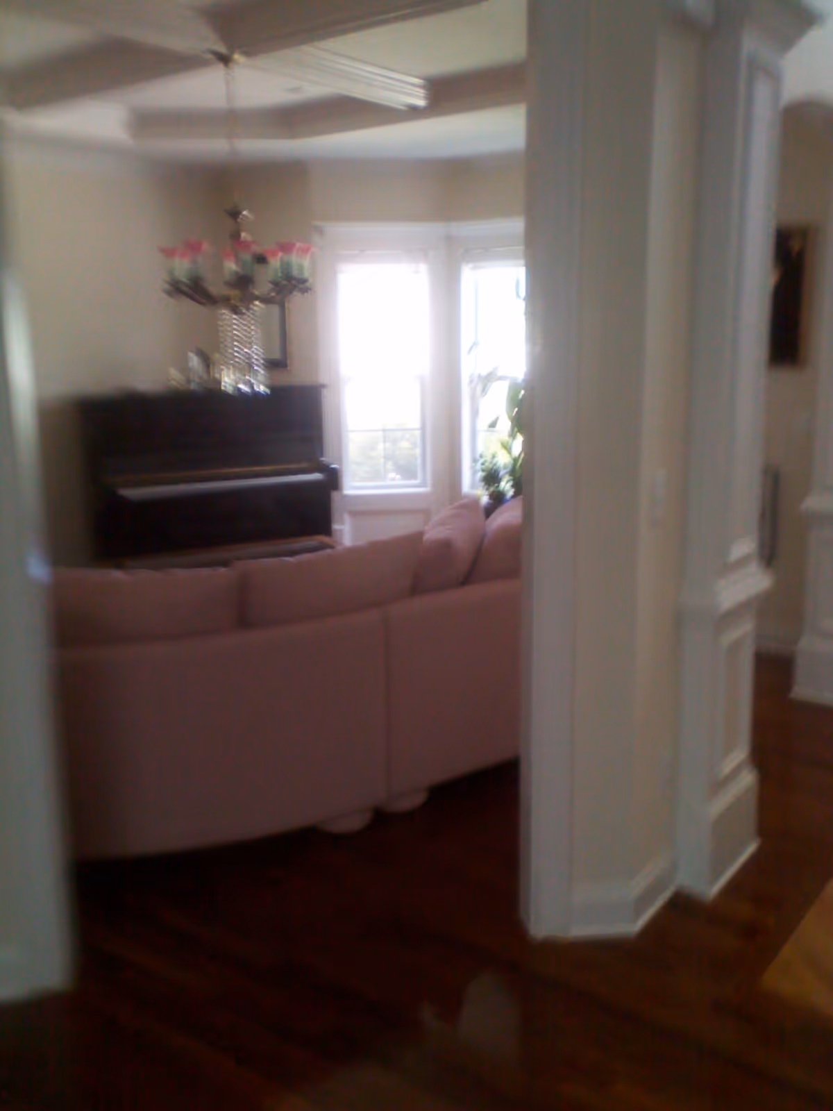 Interior view of a living room featuring a pink sectional sofa, a dark upright piano against the wall, a chandelier with pink and green shades hanging from the ceiling, and large windows letting in natural light. The room has wooden flooring and white walls with decorative molding.