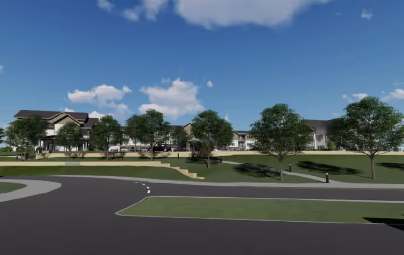 View of a large senior living facility building with multiple sections and a sloped roof, surrounded by green lawns and several trees under a blue sky with some clouds. There is a paved road and sidewalk in the foreground.