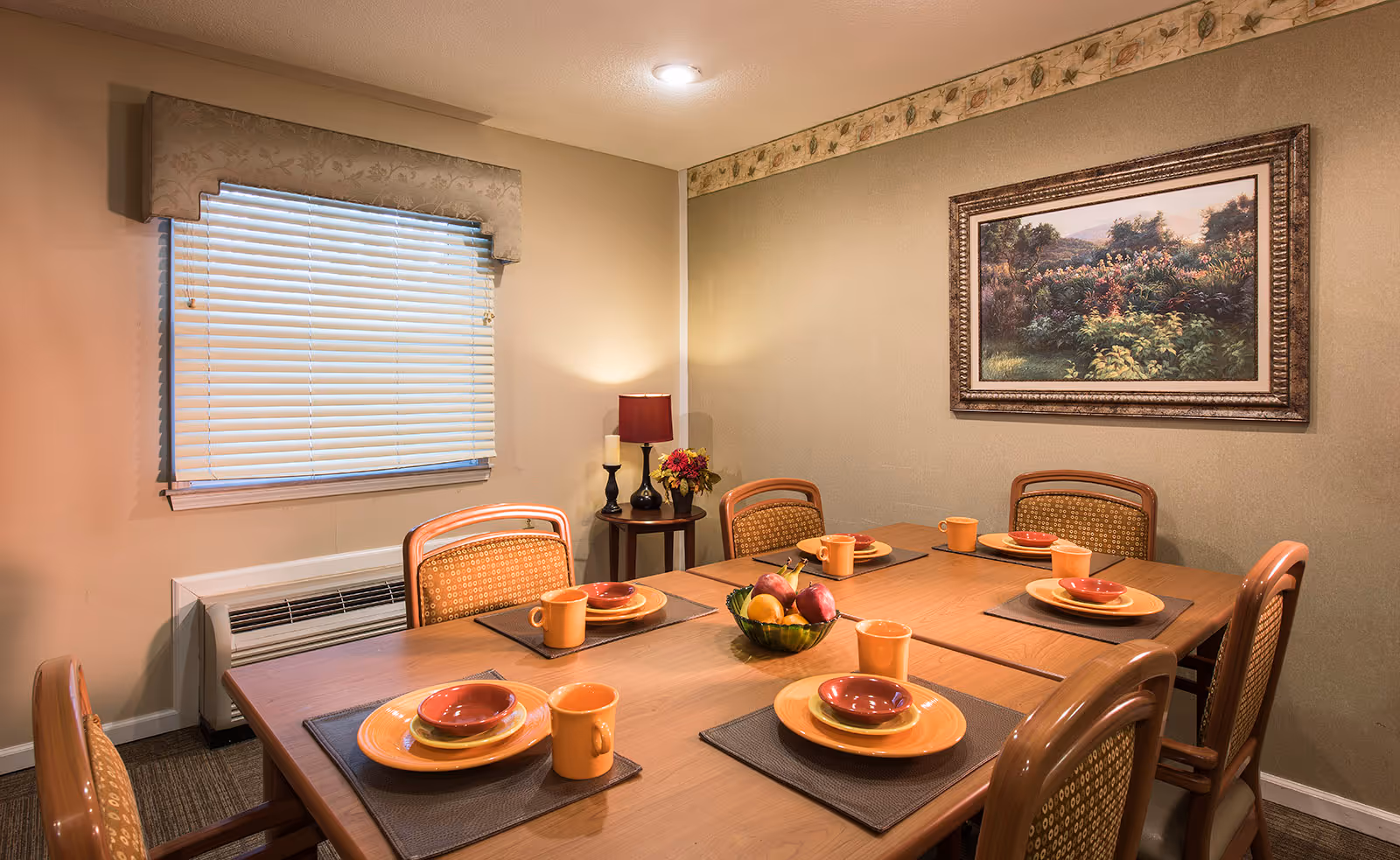 Well-lit dining room with a table set for six, plates and mugs, a fruit bowl, a framed landscape painting, and a window with blinds.
